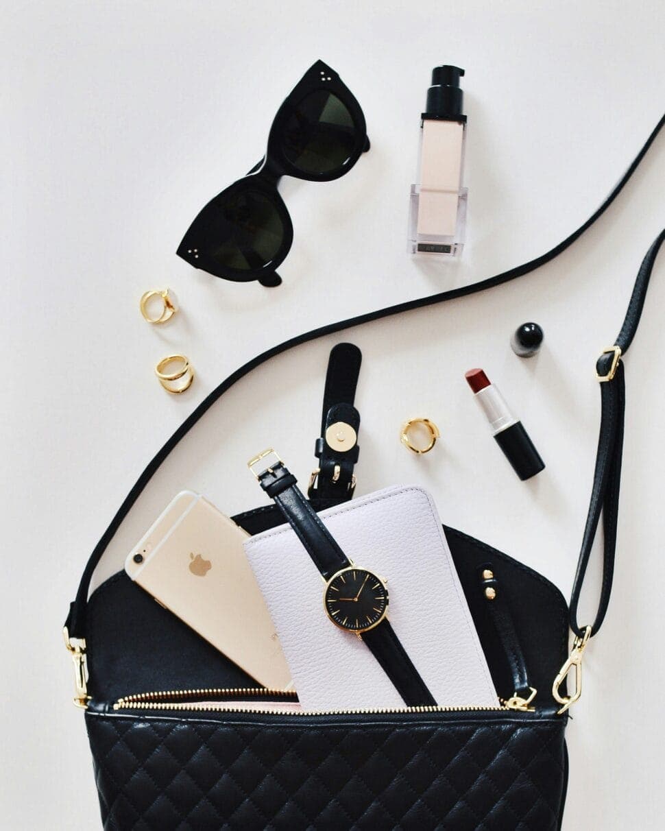 Sunglasses, a watch, lipstick, a phone and a black quilted bag laid out on white